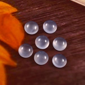 Puede incluir: Siete piedras preciosas cabuj&oacute;n transl&uacute;cidas de color azul p&aacute;lido est&aacute;n dispuestas sobre una superficie marr&oacute;n. Las piedras redondas tienen un aspecto liso y pulido, reflejando la luz. Estas piedras son adecuadas para la fabricaci&oacute;n de joyas o artesan&iacute;a.