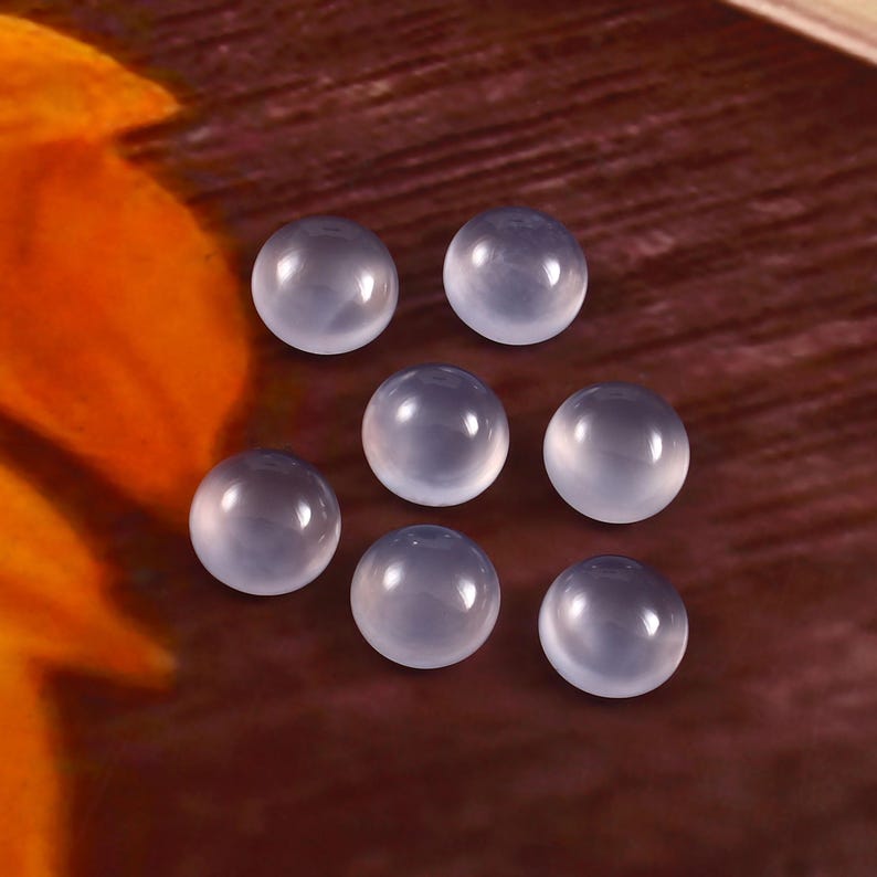 Puede incluir: Siete piedras preciosas cabuj&oacute;n transl&uacute;cidas de color azul p&aacute;lido est&aacute;n dispuestas sobre una superficie marr&oacute;n. Las piedras redondas tienen un aspecto liso y pulido, reflejando la luz. Estas piedras son adecuadas para la fabricaci&oacute;n de joyas o artesan&iacute;a.