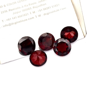 Puede incluir: Cinco gemas redondas, facetadas, de color rojo intenso están dispuestas sobre una superficie blanca. Las gemas tienen una superficie pulida y reflectante, y las facetas captan la luz. La imagen incluye texto de una tarjeta de visita.