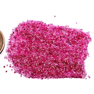 Puede incluir: Un montón de pequeñas gemas facetadas de color rosa intenso. Las gemas están dispersas sobre una superficie blanca, con algunas piedras individuales separadas del grupo principal. Las gemas son de un color rosa vibrante y saturado.