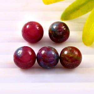 May include: Five polished gemstone beads. The beads are primarily a deep red color, with some showing patches of blue and green. The beads are round and have a smooth, glossy finish. The background is white with yellow and green leaves.