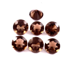 Pode incluir: Sete pedras preciosas redondas e facetadas em tons de castanho estão dispostas sobre um fundo branco. As pedras preciosas têm uma superfície polida e refletora, e as facetas criam um padrão de estrela. As pedras são provavelmente para fazer joias.