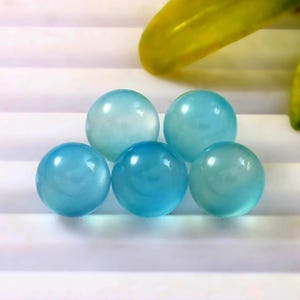May include: Five translucent, spherical, light blue gemstones. The spheres have a smooth, polished surface and varying shades of blue. The gemstones are arranged on a white surface, with a yellow leaf in the background.