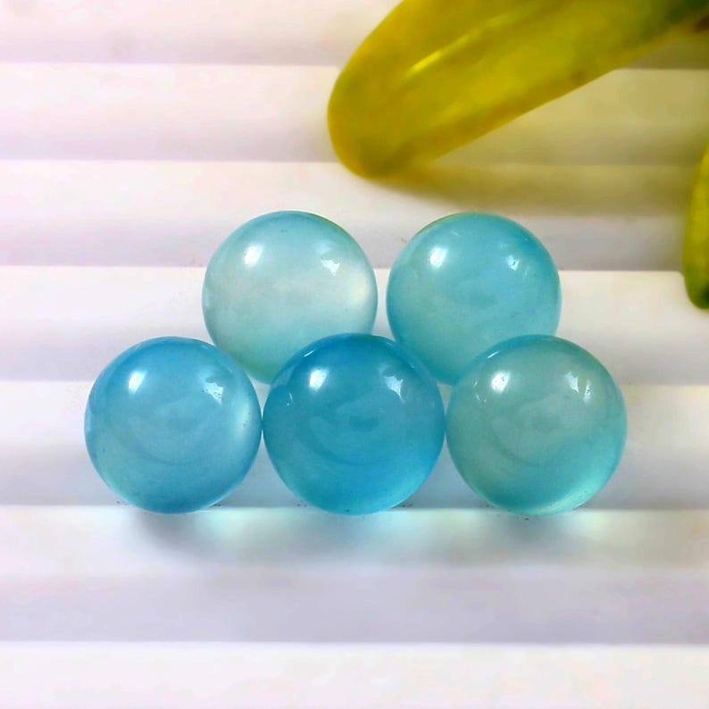 May include: Five translucent, spherical, light blue gemstones. The spheres have a smooth, polished surface and varying shades of blue. The gemstones are arranged on a white surface, with a yellow leaf in the background.