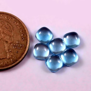 Puede incluir: Seis cabujones de piedras preciosas redondeadas de color azul claro con efecto degradado, junto a una moneda de dólar estadounidense. Las piedras preciosas tienen una superficie lisa y pulida y son una mezcla de azul claro y oscuro. La moneda es dorada.