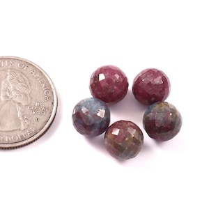 Könnte beinhalten: Fünf runde, facettierte Edelsteinperlen in den Farben Dunkelrot, Violett und Blau, angeordnet neben einer US-Vierteldollarmünze als Größenvergleich. Jede Perle hat eine facettierte Oberfläche, die das Licht reflektiert und Farbnuancen zeigt.