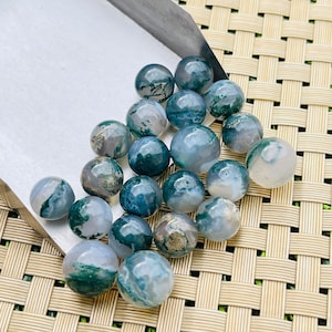 Puede incluir: Colección de cuentas redondas de ágata musgosa pulida en tonos verdes, azules y blancos. Las cuentas se exhiben en una bandeja plateada y sobre una superficie tejida de color marrón claro. Los patrones naturales crean un aspecto único.