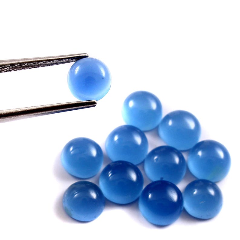 Może przedstawiać: Kolekcja dziesięciu okrągłych, wypolerowanych, jasnoniebieskich kamieni szlachetnych. Jeden kamień jest trzymany przez srebrną pęsetę. Kamienie mają jednolity rozmiar i kształt, z gładką, odblaskową powierzchnią. Tło jest białe.