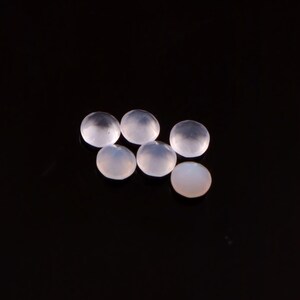 May include: Six translucent, round gemstones with a soft, milky appearance are arranged on a black surface. The gems have a smooth, polished surface, and they reflect light, creating subtle highlights and shadows.