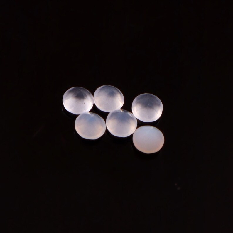May include: Six translucent, round gemstones with a soft, milky appearance are arranged on a black surface. The gems have a smooth, polished surface, and they reflect light, creating subtle highlights and shadows.