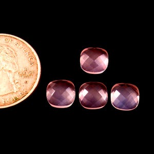 May include: Four faceted, cushion-cut, pink gemstones are displayed against a black background, with a US quarter coin for size comparison. The gemstones have a soft, translucent appearance and are arranged in a cluster.