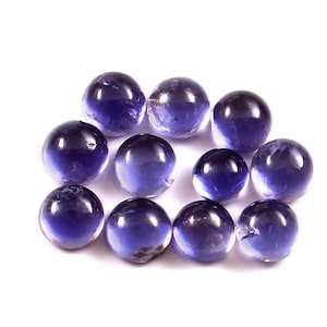 Puede incluir: Una colección de once piedras preciosas pulidas, redondas y translúcidas. Las piedras son de color violeta intenso, con una superficie vítrea y reflectante. Las piedras preciosas están dispersas sobre un fondo blanco.