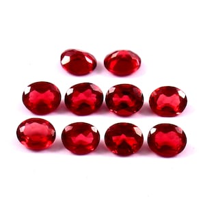 May include: A collection of ten oval-shaped, faceted, deep red gemstones. The gems are arranged on a white background, showcasing their polished surfaces and internal brilliance. The stones are similar in size and shape, suggesting they are from the same source.