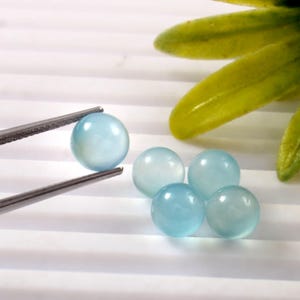 May include: A collection of light blue, spherical gemstones. One gem is held by silver tweezers, while four others are scattered nearby. The gems have a translucent quality, and the background is white with subtle lines.