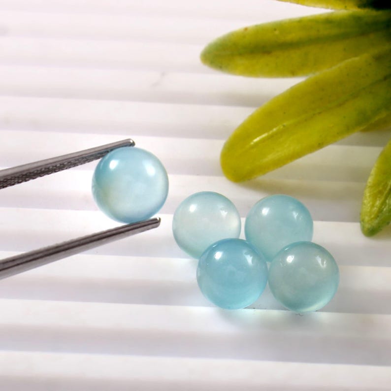 May include: A collection of light blue, spherical gemstones. One gem is held by silver tweezers, while four others are scattered nearby. The gems have a translucent quality, and the background is white with subtle lines.