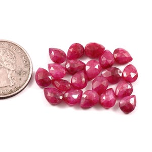 May include: A collection of faceted, deep red ruby gemstones in various shapes, including teardrop and oval, are scattered on a white surface. A US dollar coin is visible in the upper left corner, providing a size reference.