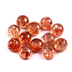 May include: Eleven polished sunstone gemstones are displayed. The round stones are a translucent orange-brown with sparkling inclusions. They are scattered on a white background. These gemstones are suitable for jewellery making.