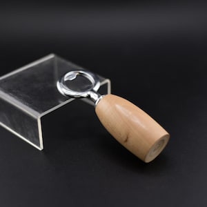 Può includere: Un apribottiglie con un manico in legno chiaro e un apribottiglie in metallo color argento. L'apribottiglie è angolato su un espositore in acrilico trasparente su sfondo nero. Il manico in legno ha una finitura liscia.