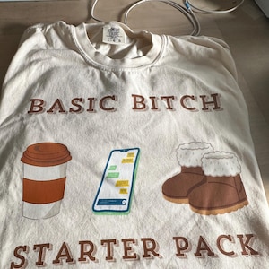 以下が含まれることがあります： ベージュのTシャツに、茶色の文字で「BASIC BITCH STARTER PACK」とプリントされています。Tシャツには、コーヒーカップ、テキストメッセージが表示されたスマートフォン、茶色のブーツの3つのイラストが描かれています。