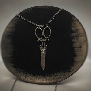 Op de afbeelding: Een zilveren ketting met een gedetailleerde schaar hanger. De ketting wordt getoond op een zwart houten oppervlak.