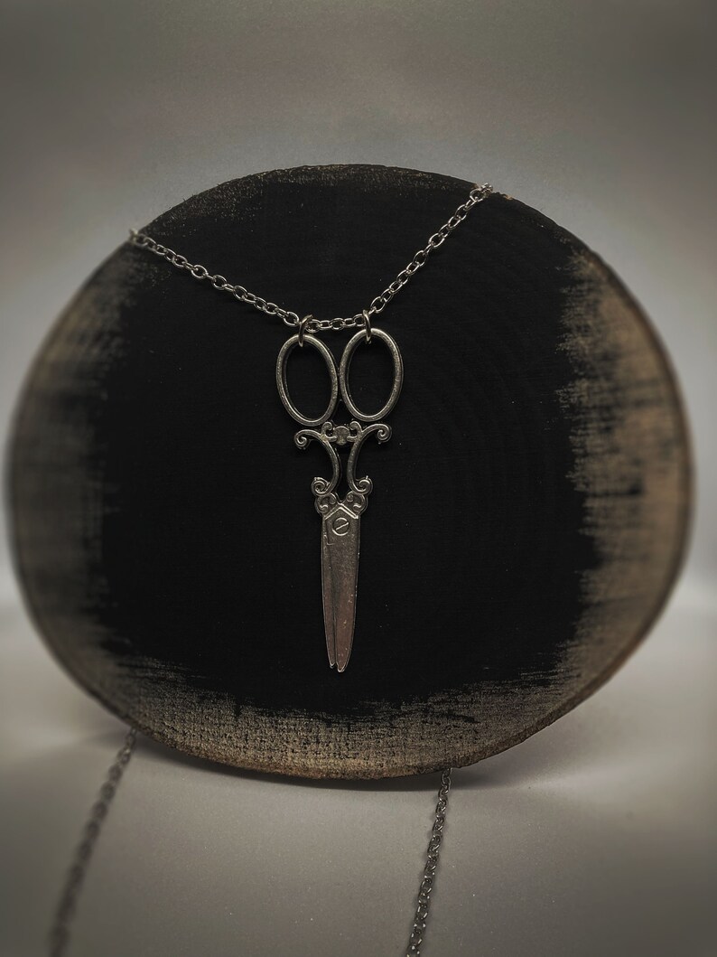 Op de afbeelding: Een zilveren ketting met een gedetailleerde schaar hanger. De ketting wordt getoond op een zwart houten oppervlak.