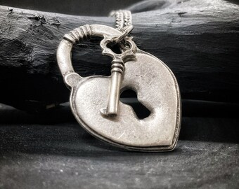 May include: A silver heart-shaped locket with a keyhole and a small key attached to it. The locket is on a silver chain and is resting on a dark surface.