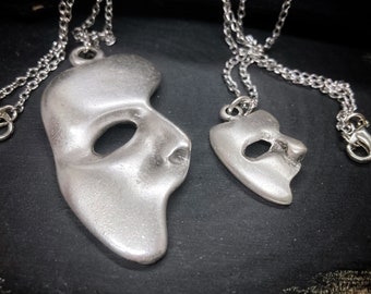 Op de afbeelding: Twee zilveren kettingen met een Phantom of the Opera-masker hanger. De grotere hanger is een gedetailleerd profiel van het masker, terwijl de kleinere hanger een meer vereenvoudigde versie is. Beide hangers zijn aan zilveren kettingen.
