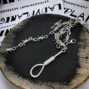 May include: A silver chain necklace with a barbed wire design and a silver noose pendant. The necklace is displayed on a black plate with the text "LADYPLANTGIFT EXTRAORDINARY ACCESSORIES" on the bottom.