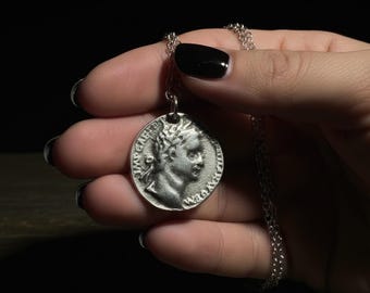 May include: Silver-toned coin pendant necklace. The pendant features a profile of a person with a laurel wreath and text around the edge. The necklace chain is delicate and silver. The background is dark.