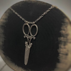 Op de afbeelding: Een zilveren ketting met een gedetailleerde, sierlijke schaar hanger. De ketting is een eenvoudige zilveren ketting.