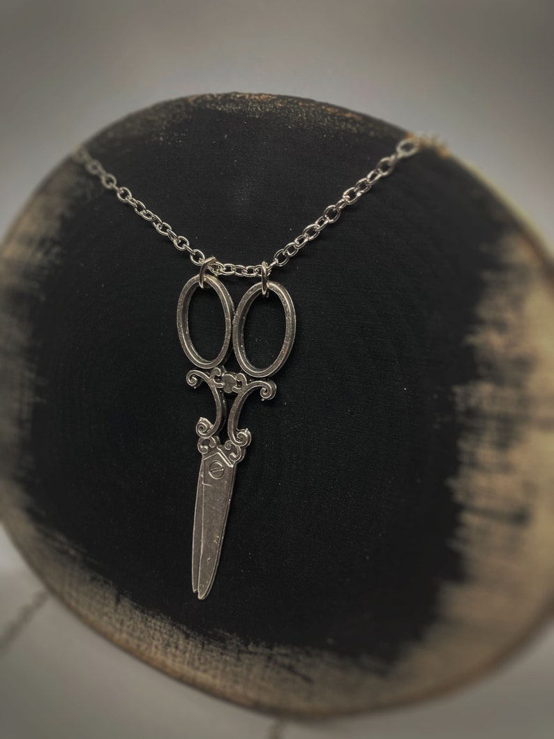 Op de afbeelding: Een zilveren ketting met een gedetailleerde, sierlijke schaar hanger. De ketting is een eenvoudige zilveren ketting.