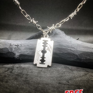 Könnte beinhalten: Silberne Halskette mit einem Rasiermesser-Anhänger und einer Stacheldrahtkette. Der Anhänger hat ein Design mit zwei Flügeln auf beiden Seiten der Klinge. Die Halskette liegt auf einer dunklen Holzoberfläche. KOSTENLOSER VERSAND.