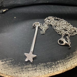 Può includere: Una collana d'argento con un ciondolo a forma di stella su una catena d'argento. Il ciondolo è a forma di bacchetta con una stella in cima.
