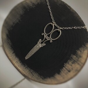 Op de afbeelding: Een zilveren schaar hanger ketting aan een zilveren ketting. De hanger is een gedetailleerd ontwerp van een schaar met een decoratieve versiering.
