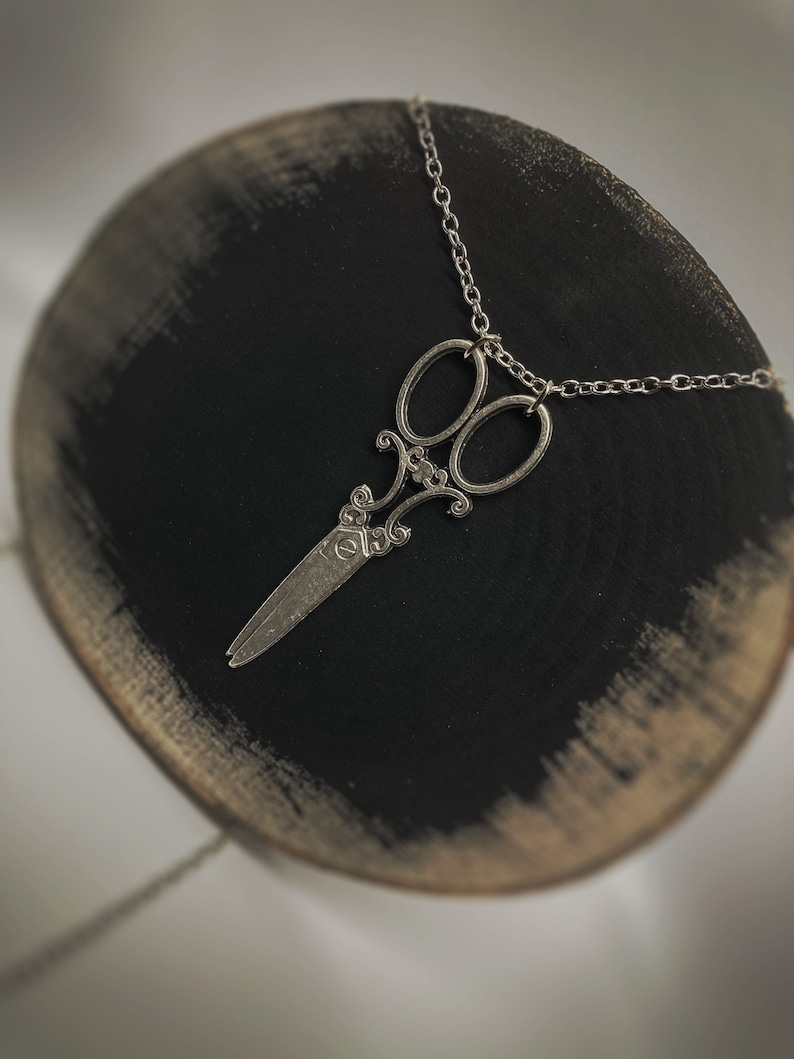 Op de afbeelding: Een zilveren schaar hanger ketting aan een zilveren ketting. De hanger is een gedetailleerd ontwerp van een schaar met een decoratieve versiering.