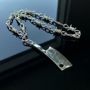 May include: A silver-colored necklace featuring a barbed wire chain and a cleaver-shaped pendant. The pendant has a small hole near the bottom. The necklace is displayed on a reflective black surface.