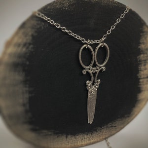 Op de afbeelding: Een zilveren ketting met een gedetailleerde, sierlijke schaar hanger. De ketting wordt getoond op een zwart en bruin houten oppervlak.