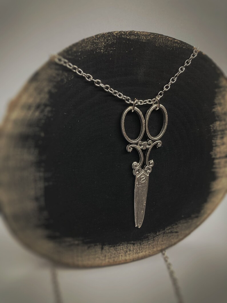 Op de afbeelding: Een zilveren ketting met een gedetailleerde, sierlijke schaar hanger. De ketting wordt getoond op een zwart en bruin houten oppervlak.