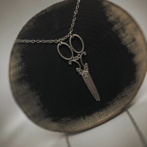 Op de afbeelding: Een zilveren ketting met een gedetailleerde, sierlijke schaar hanger. De ketting wordt gepresenteerd op een zwarte ronde standaard.