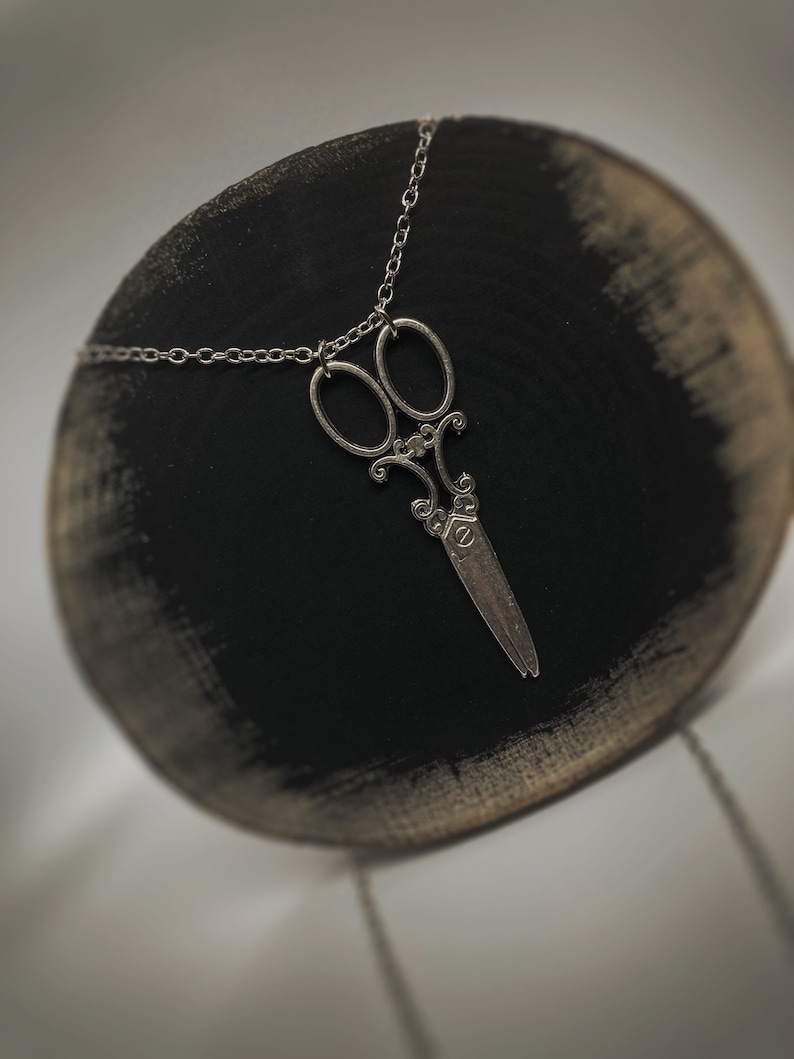 Op de afbeelding: Een zilveren ketting met een gedetailleerde, sierlijke schaar hanger. De ketting wordt gepresenteerd op een zwarte ronde standaard.