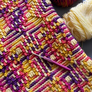 May include: A vibrant crochet piece featuring a geometric design in yellow, pink, and purple. A crochet hook lies on the fabric, with two balls of yarn in matching colours. The yarn is a mix of yellow, purple, and pink, perfect for crafting.