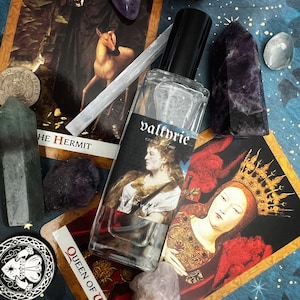 Pode incluir: Um frasco de vidro transparente de valkyrie eau de parfum com uma tampa preta, rodeado por cartas de tarô, cristais e um amuleto de prata. As cartas de tarô são O Eremita e a Rainha de Varinhas.
