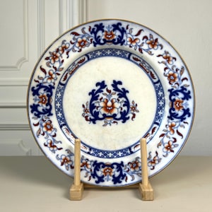 May include: A round ceramic plate featuring a white center with a central blue and orange floral motif. The rim is decorated with a repeating pattern of blue scrollwork and orange flowers, finished with a thin gold-toned edge trim.