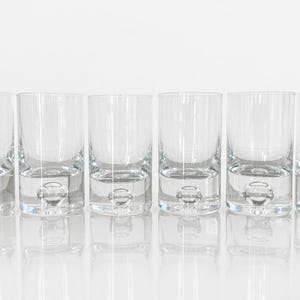 Może przedstawiać: Zestaw sześciu przezroczystych szklanek ustawionych w rzędzie. Każda cylindryczna szklanka posiada grube, solidne dno z pojedynczym, wyraźnym pęcherzykiem powietrza zatopionym w szkle, co nadaje wszystkim sześciu elementom jednolity wygląd.