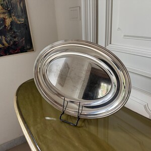 May include: A polished silver-toned oval serving tray with a smooth, reflective surface and a raised, rounded rim. The tray is displayed upright on a black wire stand, showcasing its metallic finish and simple, clean design with no ornamentation.