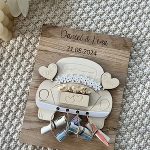 May include: A wooden wedding keepsake featuring a car design with the words "just married." The names "Daniel & Lena" and the date "21.08.2024" are engraved on the wooden plaque. Decorative elements include heart-shaped balloons and rolled-up money.