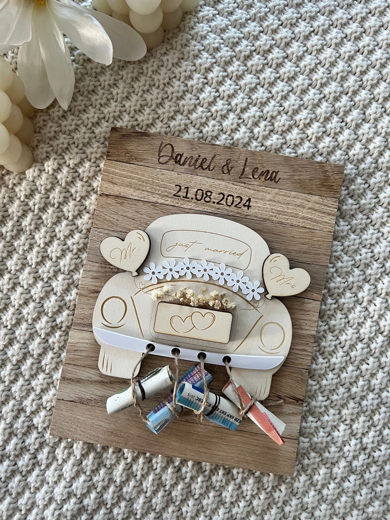 May include: A wooden wedding keepsake featuring a car design with the words "just married." The names "Daniel & Lena" and the date "21.08.2024" are engraved on the wooden plaque. Decorative elements include heart-shaped balloons and rolled-up money.