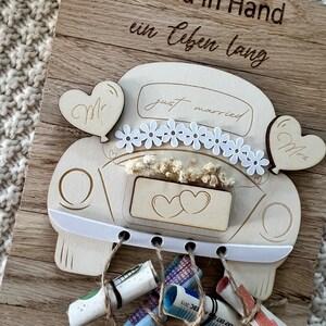 May include: Wooden wedding car decoration with the words "Hand in Hand ein Leben lang" engraved on a wooden board. The car has "just married" text, daisy accents, and heart-shaped balloons. Rolled-up money is tied to the car with twine.