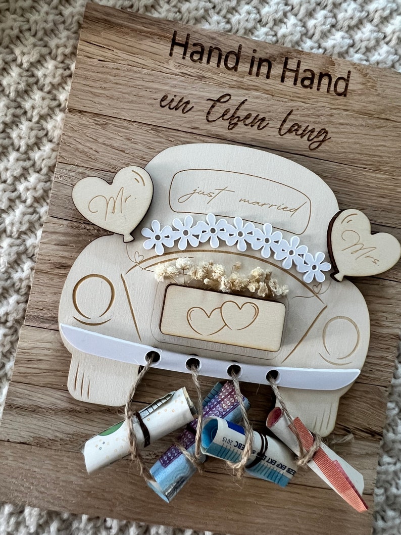 May include: Wooden wedding car decoration with the words "Hand in Hand ein Leben lang" engraved on a wooden board. The car has "just married" text, daisy accents, and heart-shaped balloons. Rolled-up money is tied to the car with twine.