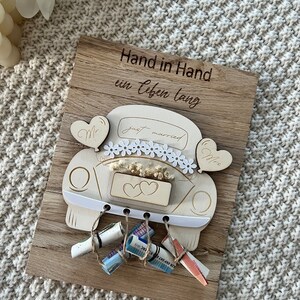 May include: A wooden wedding keepsake featuring a car design with the words "just married." The car is decorated with flowers and hearts, and the wooden plaque has the German text "Hand in Hand ein Leben lang." Several rolled-up bills are attached.